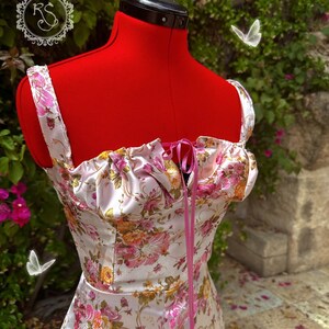 May include: A white satin dress with a floral print and pink ribbon tie. The dress has a sweetheart neckline and a fitted bodice. The dress is photographed on a red mannequin against a green leafy background.