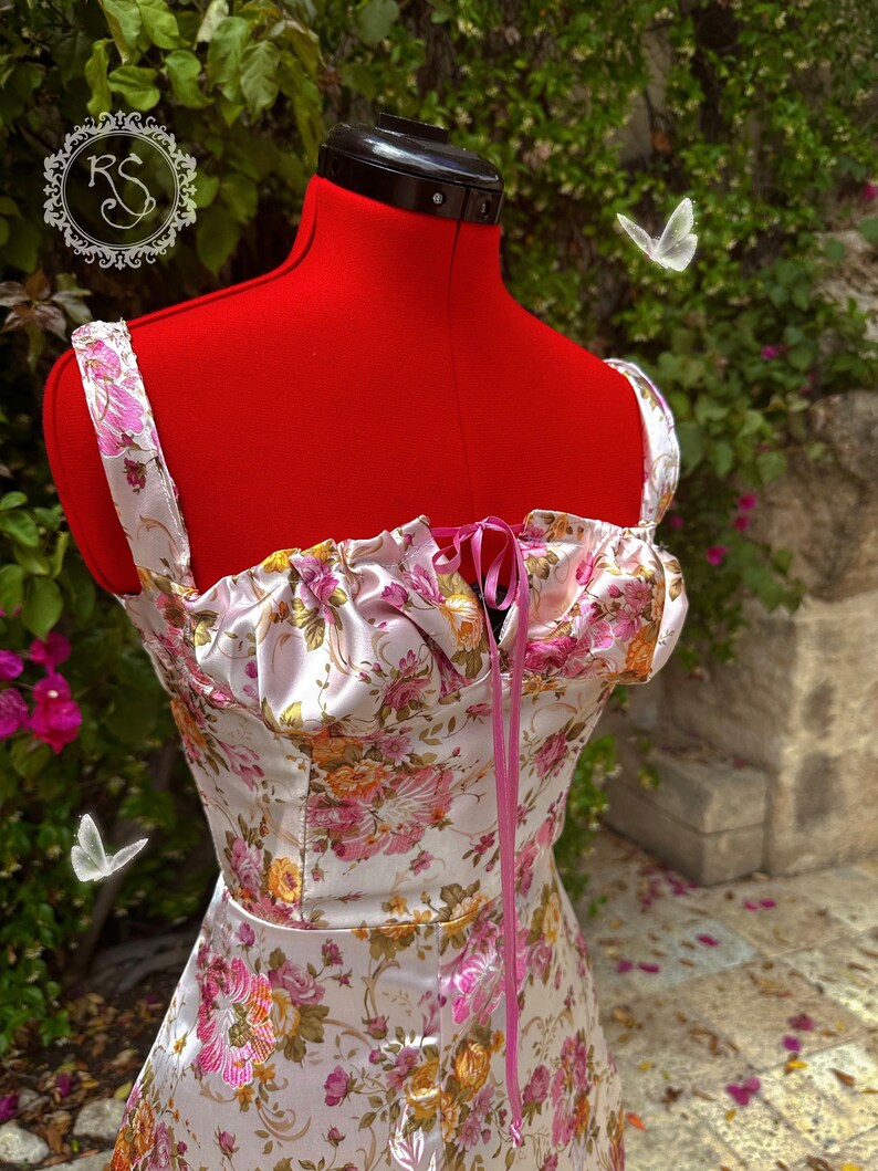 May include: A white satin dress with a floral print and pink ribbon tie. The dress has a sweetheart neckline and a fitted bodice. The dress is photographed on a red mannequin against a green leafy background.