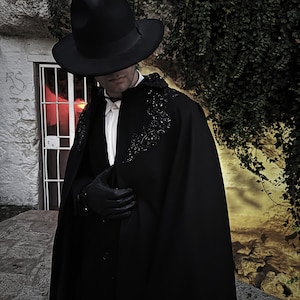 May include: A person wearing a black hat and a long black cape with beaded embellishments. The cape is worn over a white collared shirt.