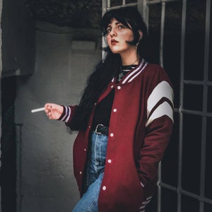 May include: A woman wearing a red varsity jacket with white stripes, blue jeans, and a black shirt. She is holding a cigarette in her right hand and looking to the right.