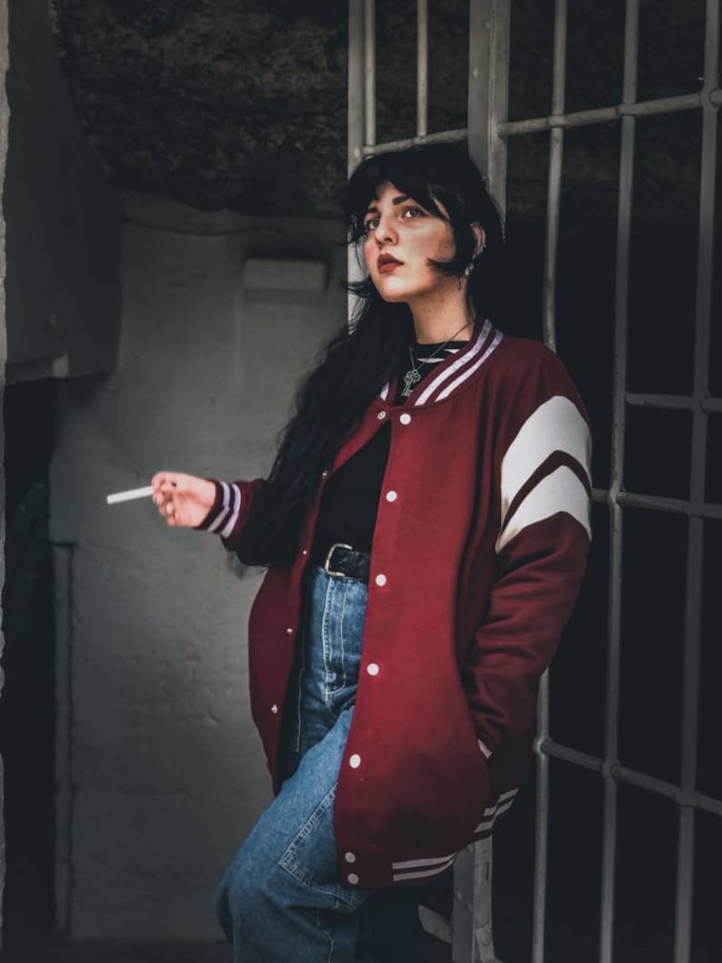 May include: A woman wearing a red varsity jacket with white stripes, blue jeans, and a black shirt. She is holding a cigarette in her right hand and looking to the right.