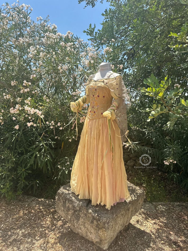 May include: A light yellow dress with a corset-style bodice adorned with floral embroidery and a flowing skirt. The dress is displayed on a mannequin atop a stone, surrounded by greenery and flowers. The dress has long sleeves and is made of a sheer fabric.