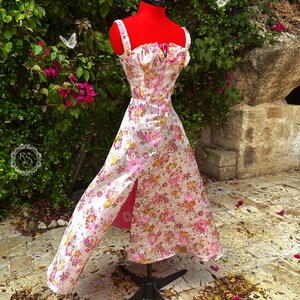 May include: A white and pink floral print dress with a pink ribbon tie at the bodice. The dress has a high neckline and a fitted bodice. The skirt is full and has a high slit on the left side. The dress is on a mannequin against a background of green foliage and stone.