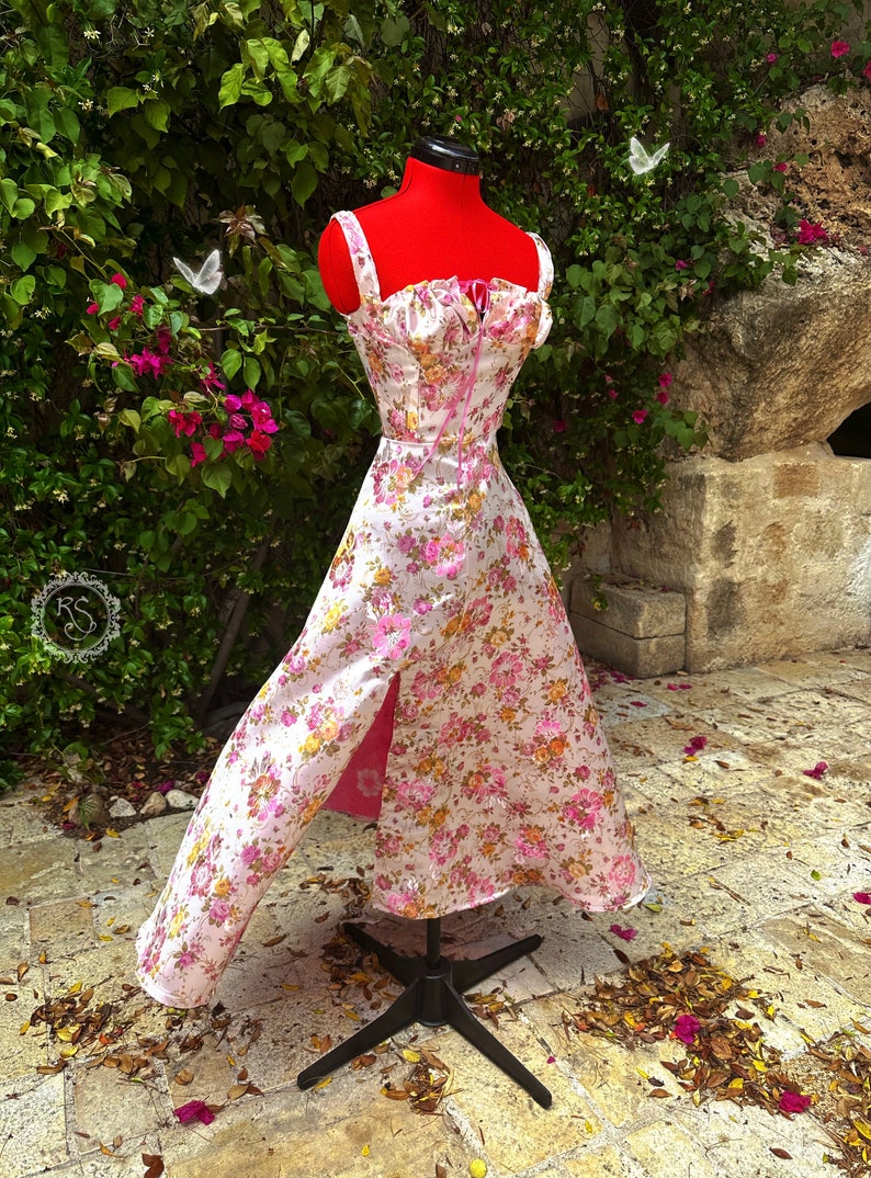 May include: A white and pink floral print dress with a pink ribbon tie at the bodice. The dress has a high neckline and a fitted bodice. The skirt is full and has a high slit on the left side. The dress is on a mannequin against a background of green foliage and stone.