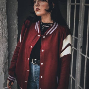 May include: A woman wearing a red and white varsity jacket with a black shirt and blue jeans. She is standing in front of a white wall and a metal door.