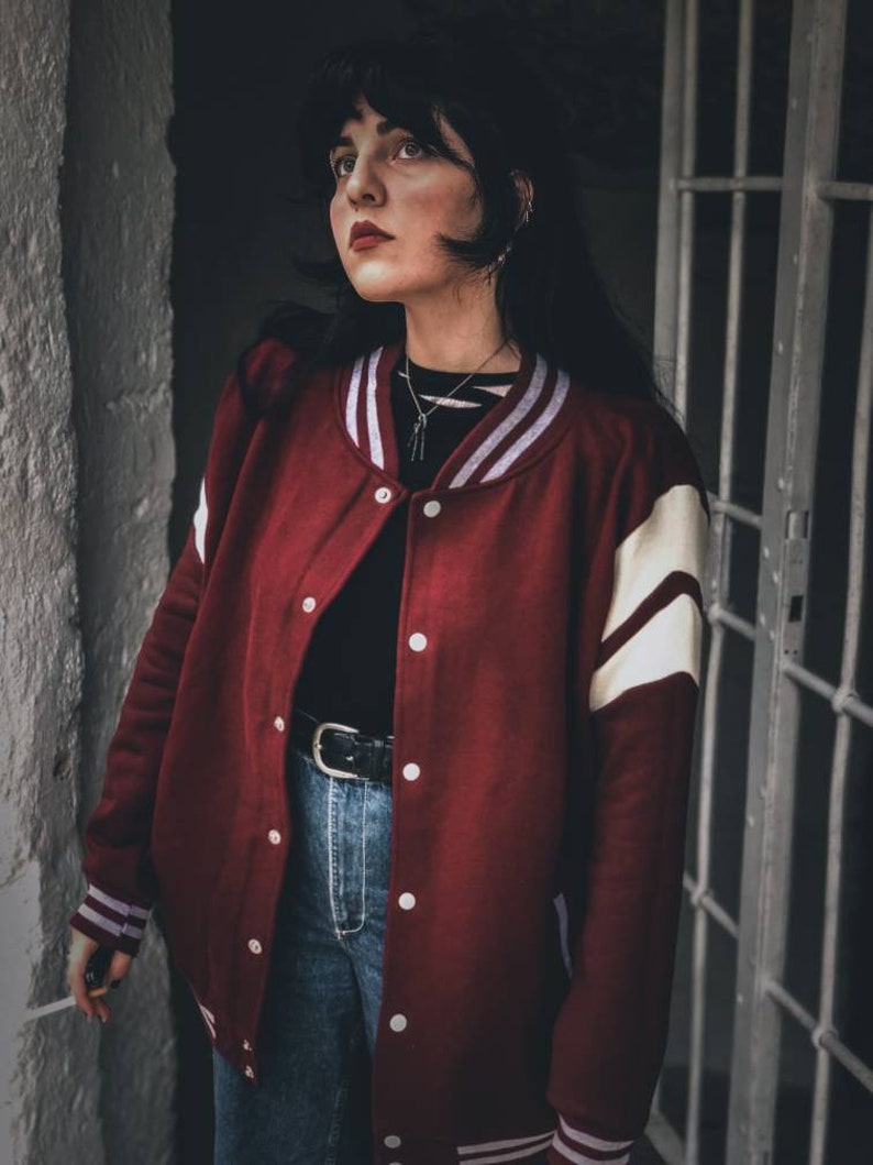 May include: A woman wearing a red and white varsity jacket with a black shirt and blue jeans. She is standing in front of a white wall and a metal door.
