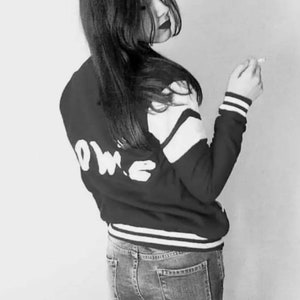 May include: A black and white photo of a person wearing a black and white varsity jacket with the word "LOVE" written on the back. The person is wearing blue jeans and has long dark hair.