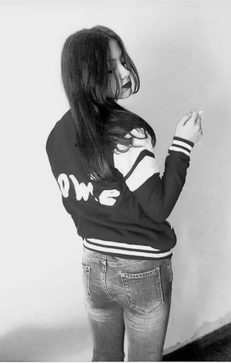 May include: A black and white photo of a person wearing a black and white varsity jacket with the word "LOVE" written on the back. The person is wearing blue jeans and has long dark hair.