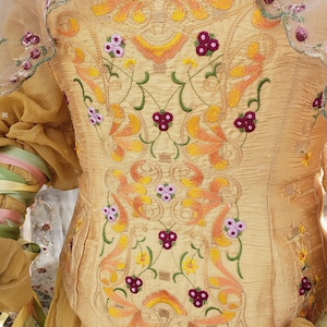 May include: A golden-yellow bodice with intricate embroidery. The embroidery features floral and leaf patterns in shades of orange, yellow, green, and purple. The bodice has a fitted shape and is made of a textured fabric. The sleeves are sheer and adorned with floral accents.