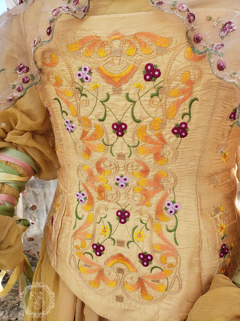 May include: A golden-yellow bodice with intricate embroidery. The embroidery features floral and leaf patterns in shades of orange, yellow, green, and purple. The bodice has a fitted shape and is made of a textured fabric. The sleeves are sheer and adorned with floral accents.