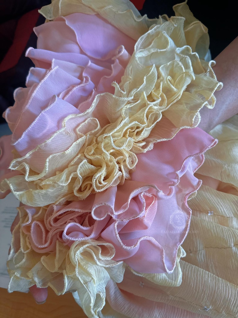 May include: Close-up of a layered fabric design with ruffled edges. The fabric features a combination of pale pink and cream colors. The ruffles create a textured, voluminous effect, suggesting a delicate and ornate textile.