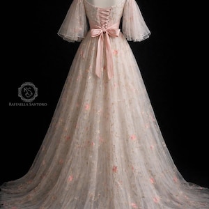 May include: An ivory, off-the-shoulder gown with pink floral embroidery. The dress has sheer, bell sleeves and a pink satin ribbon that cinches the waist. The back features a corset-style closure. Suitable for special occasions, the gown falls to the floor.