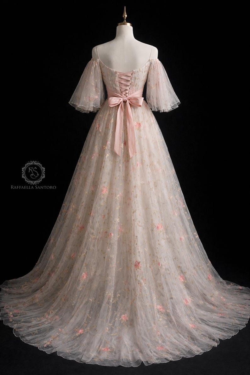 May include: An ivory, off-the-shoulder gown with pink floral embroidery. The dress has sheer, bell sleeves and a pink satin ribbon that cinches the waist. The back features a corset-style closure. Suitable for special occasions, the gown falls to the floor.