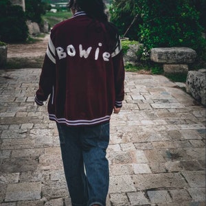 May include: A person wearing a maroon bomber jacket with white lettering that says "BOWIE" on the back. The person is wearing blue jeans and walking on a stone path.