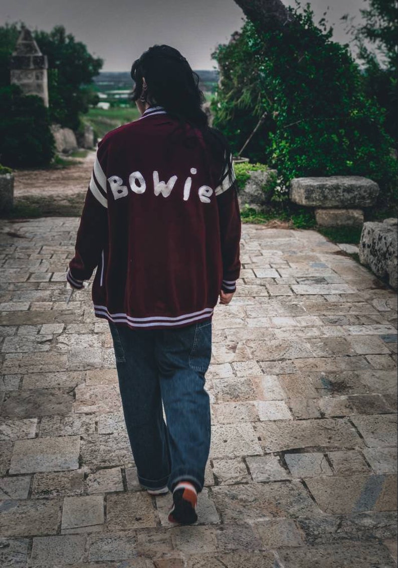 May include: A person wearing a maroon bomber jacket with white lettering that says "BOWIE" on the back. The person is wearing blue jeans and walking on a stone path.
