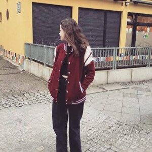 May include: A woman wearing a maroon varsity jacket with white stripes, black pants, and white sneakers. The jacket has a zipper and a button closure.
