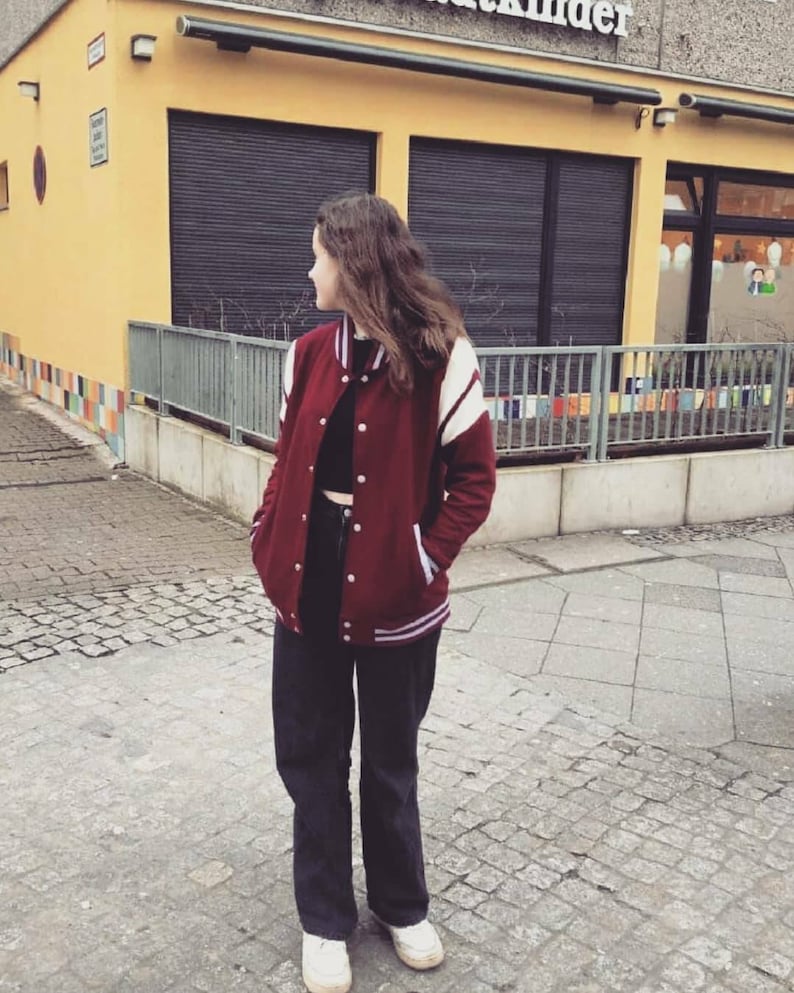 May include: A woman wearing a maroon varsity jacket with white stripes, black pants, and white sneakers. The jacket has a zipper and a button closure.