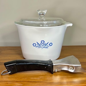 May include: A white casserole dish with a blue floral pattern and a clear glass lid, accompanied by a detachable metal handle with a black plastic grip. The dish features a flared rim and a small side tab, while the handle has a silver finish.