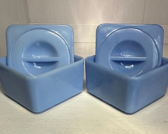 May include: Two light blue square ceramic containers with lids. The lids have a raised, circular design with a small, rectangular cutout in the center.