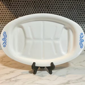 May include: A white oval serving platter with a raised center section and blue floral accents on each end. The platter is supported by a black stand and is displayed on a marble surface.