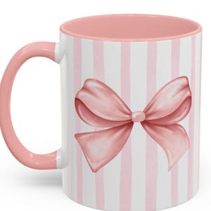 May include: A white ceramic mug with a pink interior and handle. The exterior features a vertical striped pattern in white and light pink, with a large pink bow in the center. The mug is designed for hot beverages.