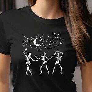 May include: Black t-shirt featuring a white graphic of three dancing skeletons under a crescent moon and stars. The skeletons are in various dance poses, with their arms and legs outstretched. The design is centered on the shirt.