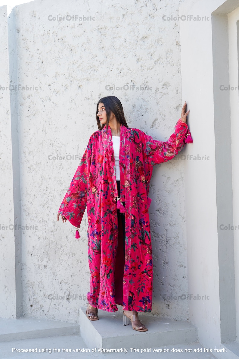 May include: A long, pink velvet robe with a floral print. The robe has a shawl collar and long sleeves. The robe is worn by a person standing in front of a white wall.