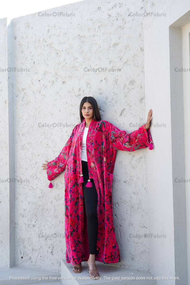 May include: A long, pink velvet robe with a floral print and tassels. The robe is worn by a person standing in front of a white wall.