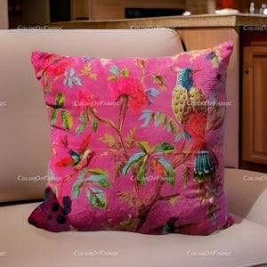 May include: Pink velvet throw pillow with a floral and bird print. The pillow features a large bird with blue, green, and red feathers, and smaller birds with red, green, and blue feathers. The background is a pink floral pattern.