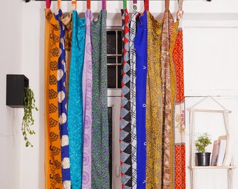Op de afbeelding: Een set kleurrijke gordijnen gemaakt van gerecyclede sari's. De gordijnen hangen aan een bruine gordijnroede en hebben verschillende patronen en kleuren, waaronder oranje, blauw, groen, paars en rood.