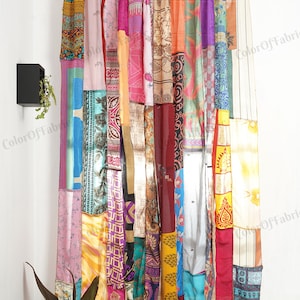 Op de afbeelding: Een kleurrijk patchwork-gordijn gemaakt van verschillende stoffen met verschillende patronen en kleuren. Het gordijn hangt aan een witte muur met een zilveren gordijnroede.