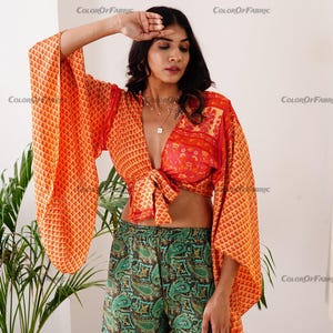 May include: A woman wearing a bright orange and red patterned crop top with a tie front and wide bell sleeves. She is also wearing green paisley patterned shorts.