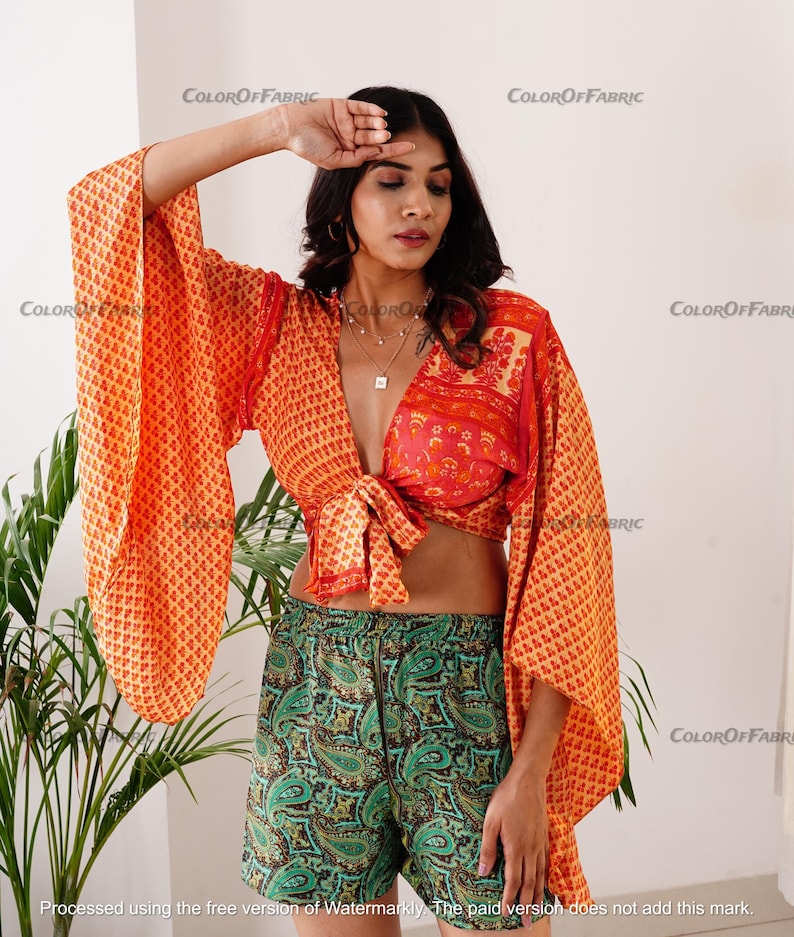 May include: A woman wearing a bright orange and red patterned crop top with a tie front and wide bell sleeves. She is also wearing green paisley patterned shorts.