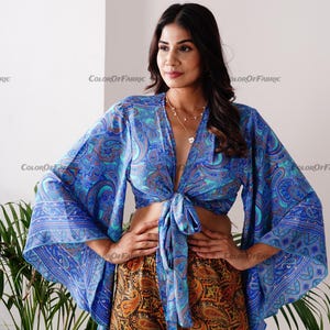 May include: A blue and turquoise paisley print crop top with long bell sleeves. The top is tied at the front and has a deep V-neckline.
