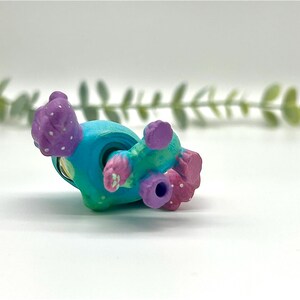Peut inclure: Une figurine miniature peinte &agrave; la main, dans des tons de turquoise, de bleu sarcelle et de violet. La figurine a un corps arrondi avec des d&eacute;tails textur&eacute;s et de petites protub&eacute;rances arrondies. Des points blancs ornent les zones violettes. La figurine est couch&eacute;e sur le c&ocirc;t&eacute;.