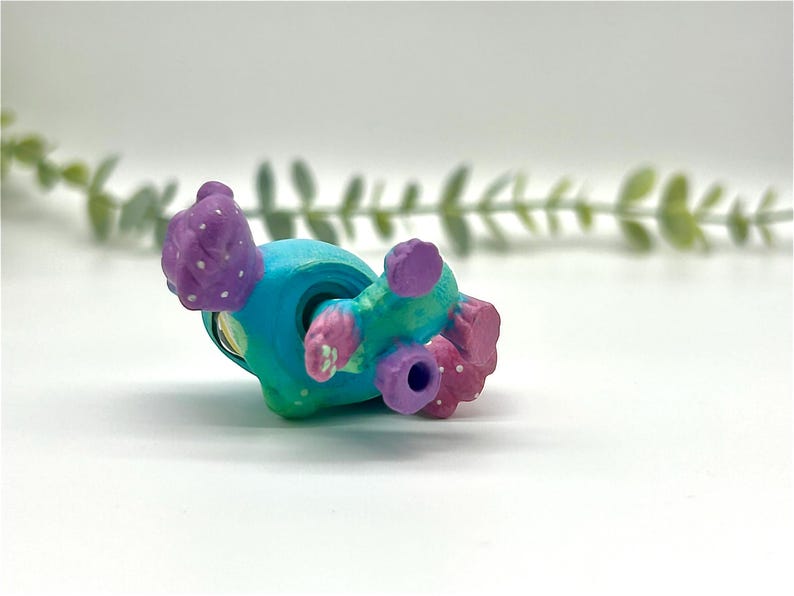 Peut inclure: Une figurine miniature peinte &agrave; la main, dans des tons de turquoise, de bleu sarcelle et de violet. La figurine a un corps arrondi avec des d&eacute;tails textur&eacute;s et de petites protub&eacute;rances arrondies. Des points blancs ornent les zones violettes. La figurine est couch&eacute;e sur le c&ocirc;t&eacute;.