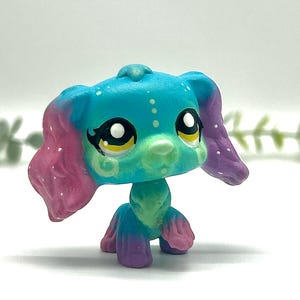 Peut inclure: Figurine de chien jouet color&eacute;e avec de grands yeux expressifs. Le chien a un corps turquoise, des oreilles roses et violettes et un museau vert. Des points et des tourbillons blancs d&eacute;corent la figurine, pos&eacute;e sur une surface blanche.