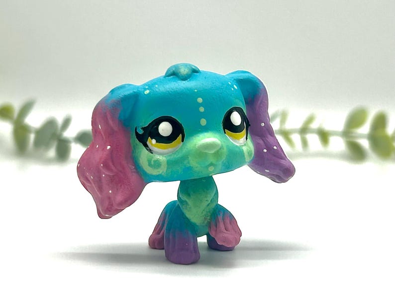 Peut inclure: Figurine de chien jouet color&eacute;e avec de grands yeux expressifs. Le chien a un corps turquoise, des oreilles roses et violettes et un museau vert. Des points et des tourbillons blancs d&eacute;corent la figurine, pos&eacute;e sur une surface blanche.
