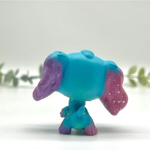 Peut inclure: Petite figurine turquoise repr&eacute;sentant un animal aux grandes oreilles. Les oreilles sont peintes en violet avec des mouchetures blanches et les pointes sont roses. La figurine a une petite queue.