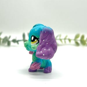 Peut inclure: Figurine de chien stylis&eacute;e et color&eacute;e, avec une t&ecirc;te bleue, un visage vert et une oreille violette. Le chien a un n&oelig;ud rose et des pois blancs sur l'oreille. Les yeux sont grands et expressifs.