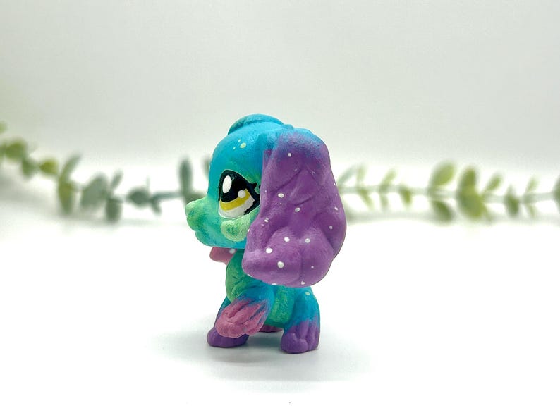 Peut inclure: Figurine de chien stylis&eacute;e et color&eacute;e, avec une t&ecirc;te bleue, un visage vert et une oreille violette. Le chien a un n&oelig;ud rose et des pois blancs sur l'oreille. Les yeux sont grands et expressifs.