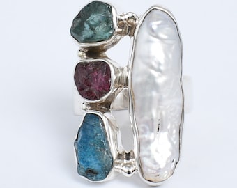 May include: A silver ring featuring three rough-cut gemstones in teal, magenta, and blue, alongside a large, elongated white pearl. The ring has a unique, open design with the gemstones set in individual bezels.