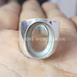 May include: A silver-colored ring with an oval-shaped setting. The ring is thick and has a polished finish. The text "THE GEMS WORLD INDIA" is printed on the ring. This ring is designed to hold a gemstone.