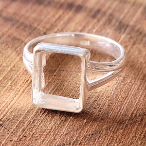 Pode incluir: Um anel de prata com uma moldura retangular. O anel tem um design de haste dividida e é colocado em uma superfície de madeira. O anel provavelmente foi projetado para conter uma pedra preciosa ou outro elemento decorativo.