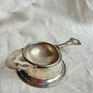 May include: A silver tea strainer with a perforated bowl and decorative handle. The strainer rests on a small, round base with an ornate, openwork design. The handle features a floral design.