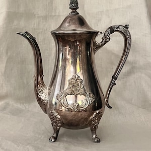May include: A silver coffee pot with intricate detailing. The pot has a curved spout, a decorative handle, and a lid with a finial. The body features an embossed design and rests on three small feet. A vintage kitchenware item.