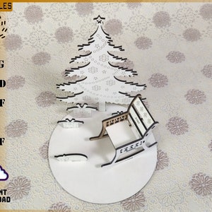 May include: A white laser-cut Christmas scene featuring a tree, presents, and a rocking chair on a circular base. The tree has snowflake and star cutouts. The image includes the text "LACUFILES" and "INSTANT DOWNLOAD".