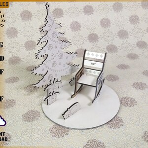May include: A laser-cut wooden Christmas scene featuring a tree, rocking chair, and sleeping cat on a circular base. The chair back displays "2021". The tree is decorated with stars and ornaments. The scene is set on a patterned background.