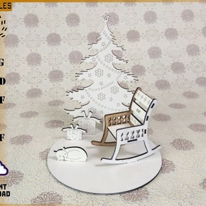 May include: A laser-cut Christmas scene featuring a tree, presents, a rocking chair, and a sleeping cat. The chair has a brown seat and white frame. The scene is on a white circular base. The text "INSTANT DOWNLOAD" is visible.