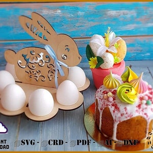 Könnte beinhalten: Ostereierhalter aus Holz in Form eines Hasen mit sechs weißen Eiern. Der Hase ist mit einem blauen Band und Blumenmustern verziert. Im Hintergrund ein dekorierter Osterkuchen mit weißem Zuckerguss und bunten Baiser-Dekorationen.
